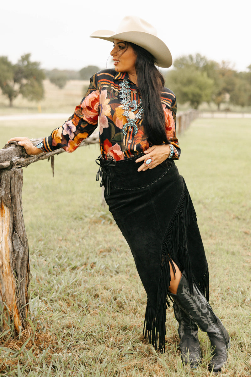 Woman in a cowboy hat and patterned shirt standing in a field with a wooden post.