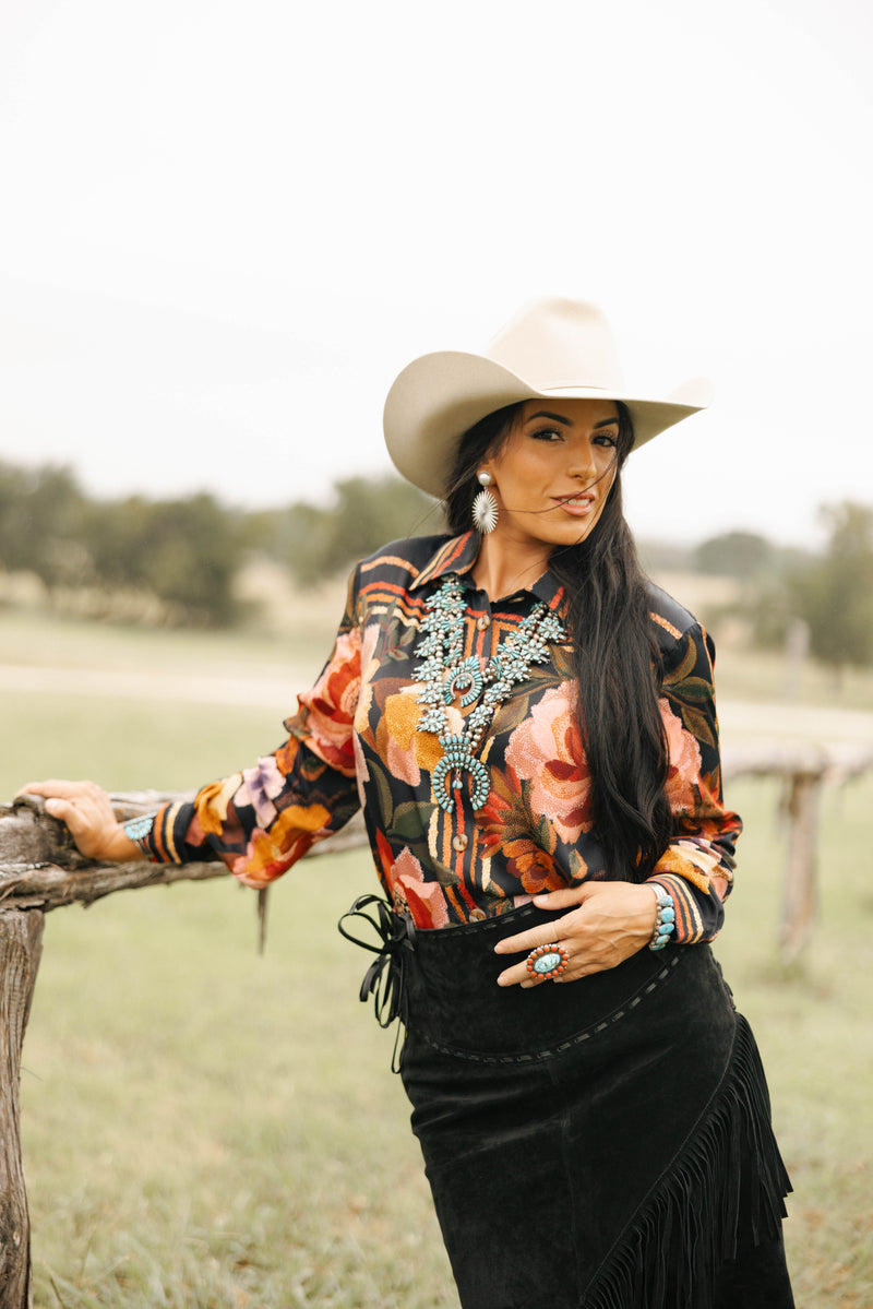 Woman in a colorful blouse and black skirt with a cowboy hat, standing in a field.