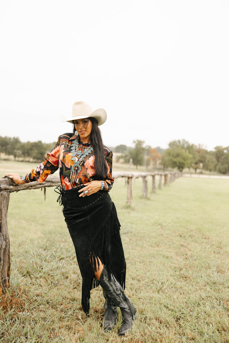 Woman in a cowboy hat and patterned shirt leaning against a wooden fence in a grassy field.