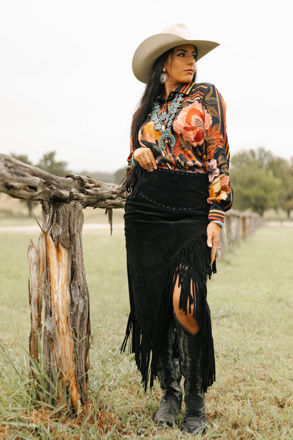 Woman in a floral blouse and black skirt standing near a wooden fence in a field.