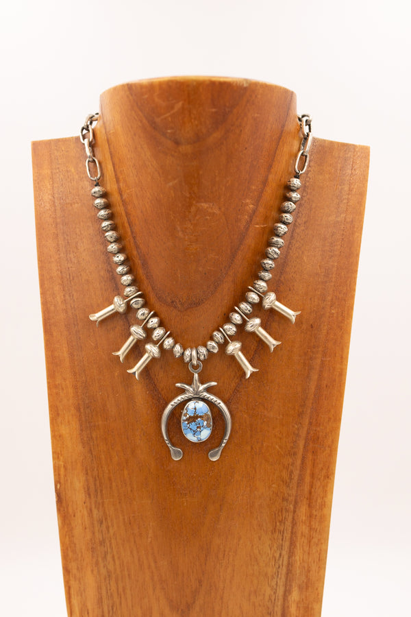 Mini squash blossom with sterling silver beads, blossoms and Naja center pendant. In the middle of the Naja pendant is a stunning light blue turquoise stone. This necklace is displayed on a wooden neck.