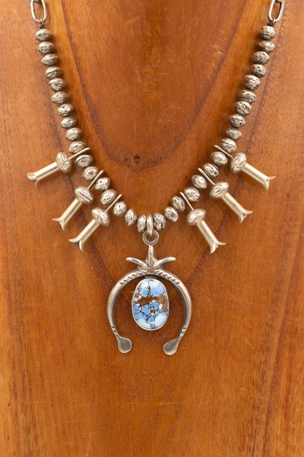 Mini squash blossom with sterling silver beads, blossoms and Naja center pendant. In the middle of the Naja pendant is a stunning light blue turquoise stone. This necklace is displayed on a wooden neck.