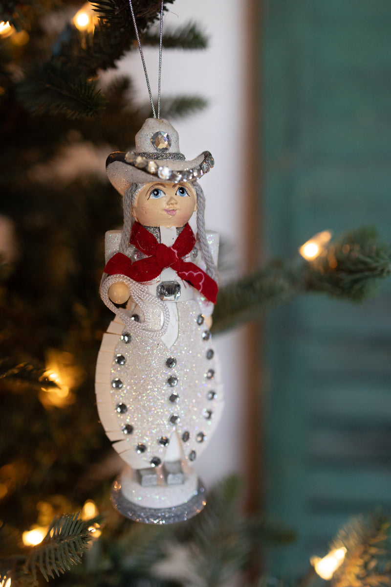 WOODEN COWGIRL CHRISTMAS ORNAMENT HANDING OFF OF A CHRISTMAS TREE. SHE HAS RHINESTONE EMBELISHMENTS.