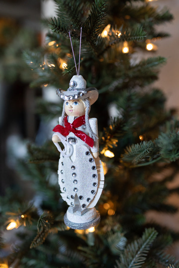 WOODEN COWGIRL CHRISTMAS ORNAMENT HANDING OFF OF A CHRISTMAS TREE. SHE HAS RHINESTONE EMBELISHMENTS.