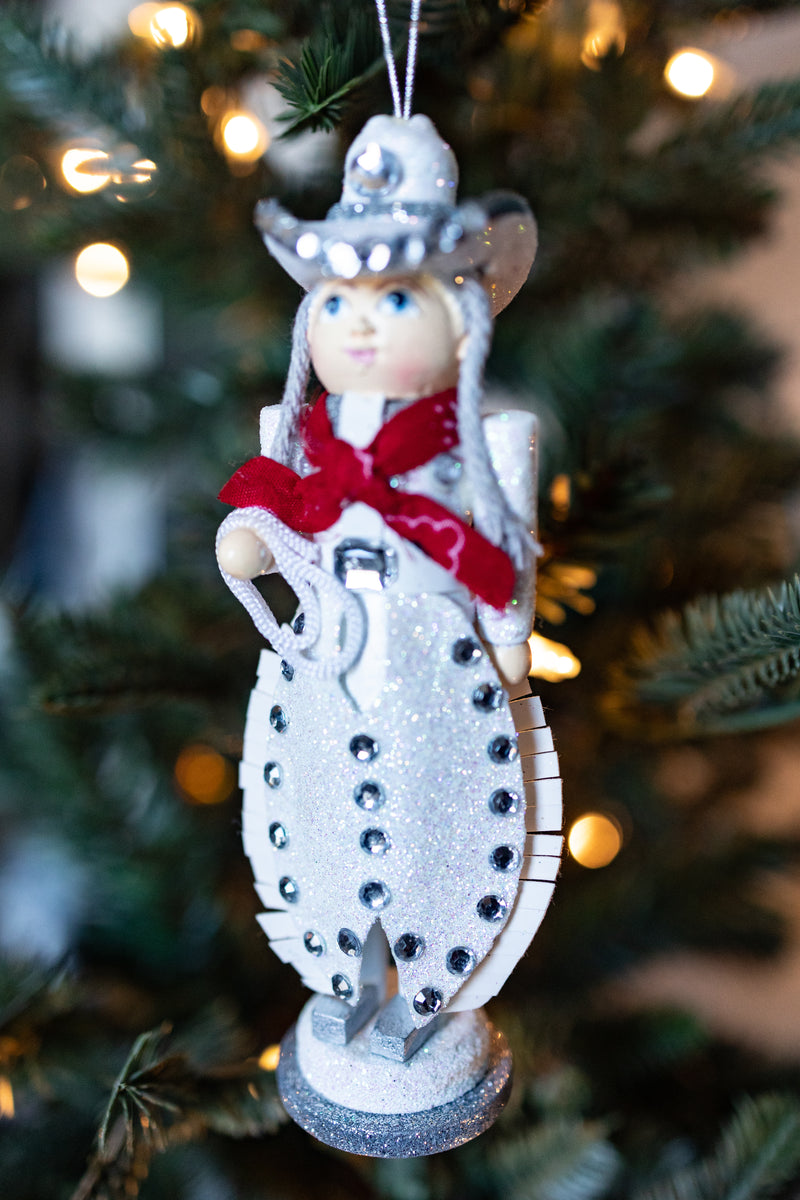 WOODEN COWGIRL CHRISTMAS ORNAMENT HANDING OFF OF A CHRISTMAS TREE. SHE HAS RHINESTONE EMBELISHMENTS.