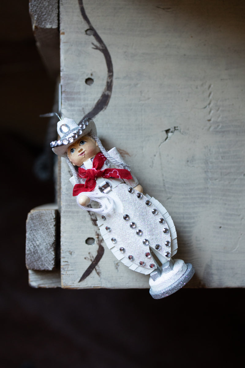 WOODEN COWGIRL CHRISTMAS ORNAMENT HANDING OFF OF A CHRISTMAS TREE. SHE HAS RHINESTONE EMBELISHMENTS.