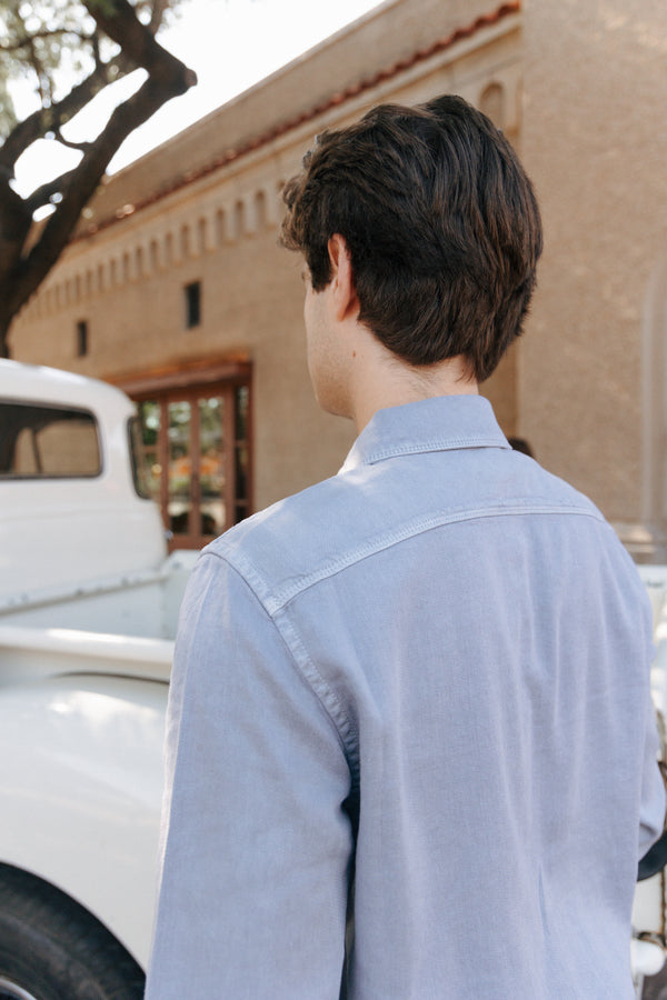 Person wearing a light blue shirt standing in front of a white truck and beige building.