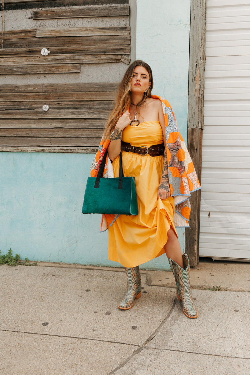 Woman in a yellow dress with a green bag standing against a blue wall.