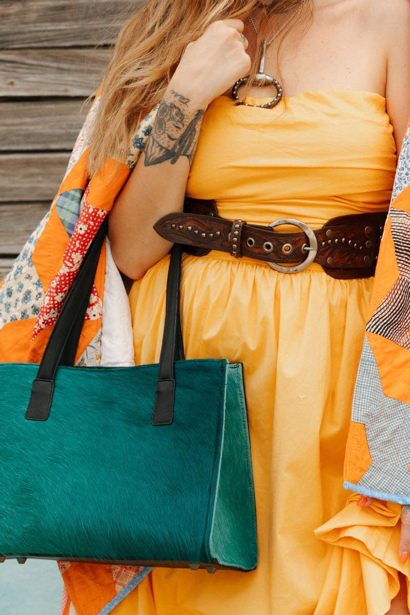 Person wearing a yellow dress with a brown belt, holding a green handbag against a wooden background.
