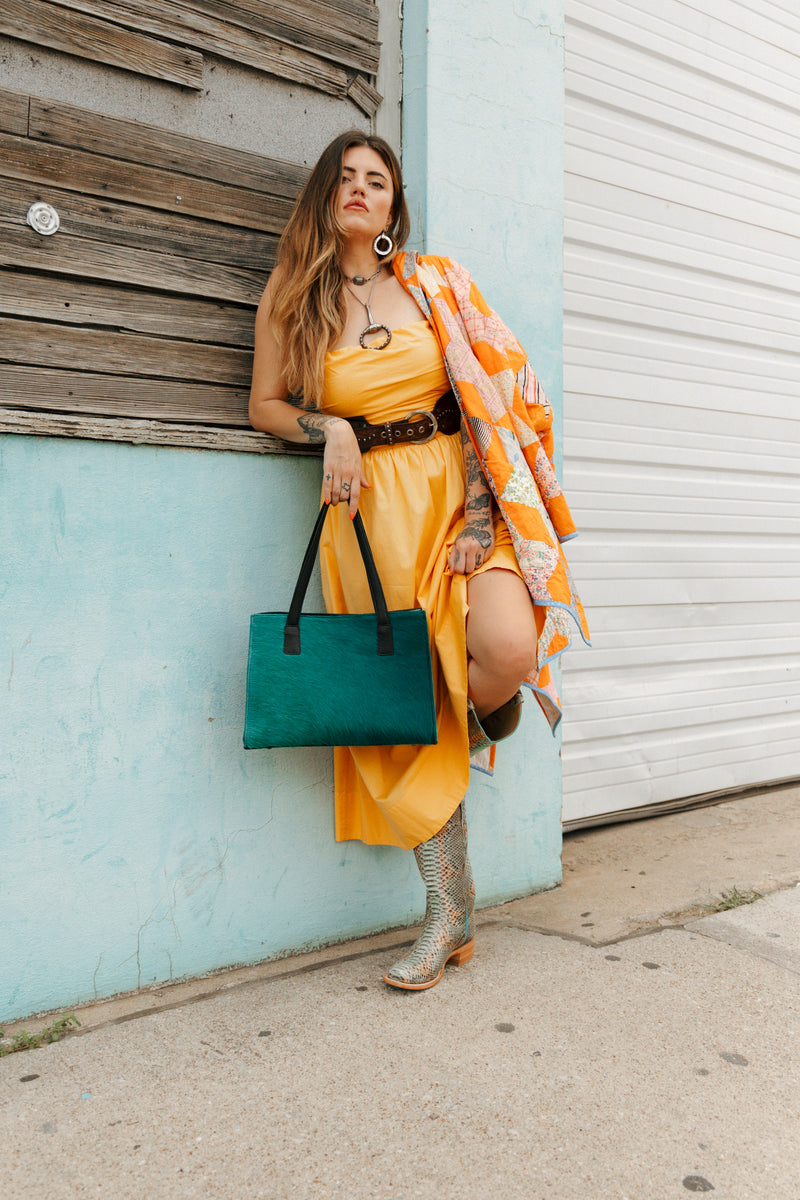 Woman in a yellow dress with a green bag leaning against a light blue wall.