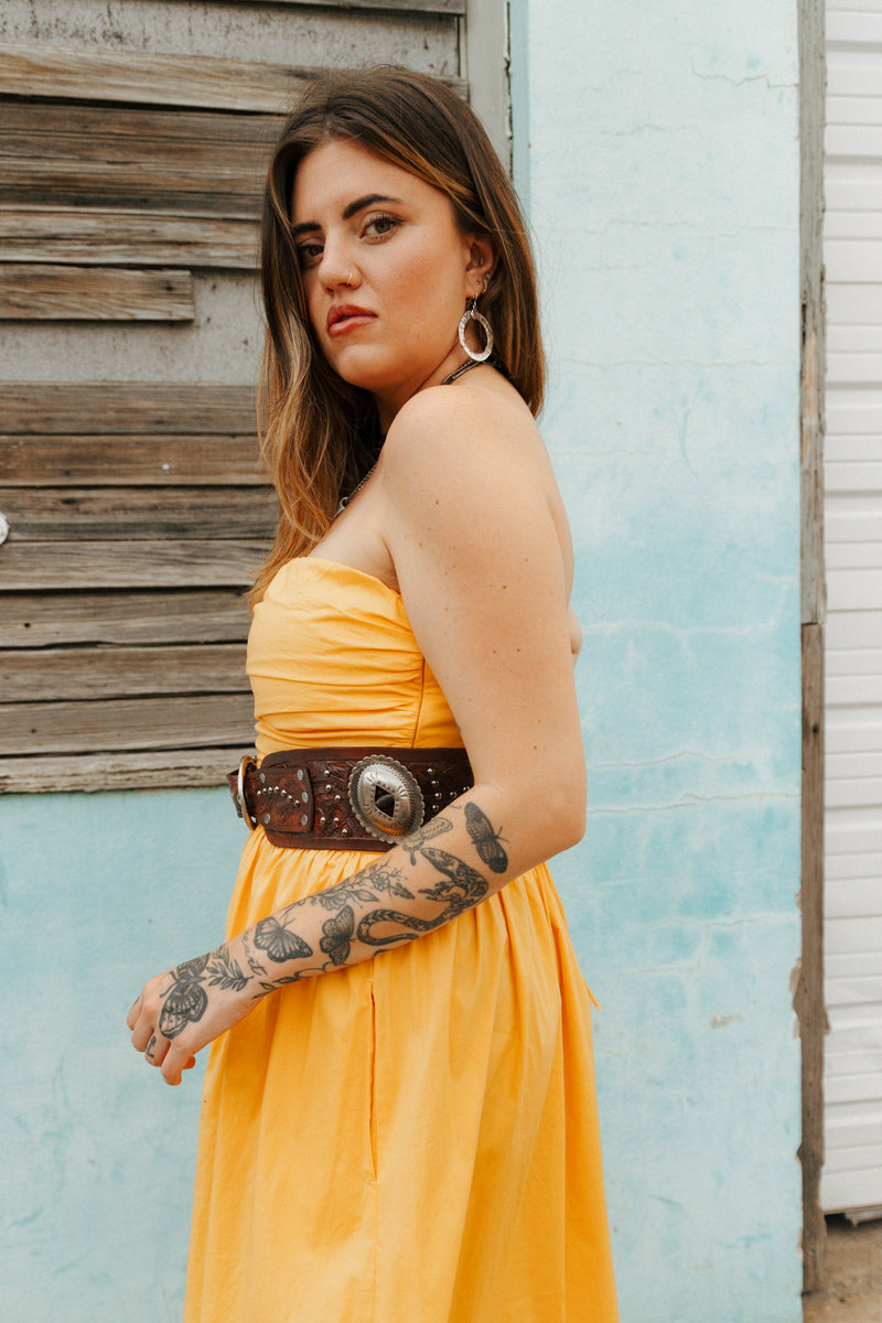 Woman in a yellow dress with a brown belt standing against a light blue wall.