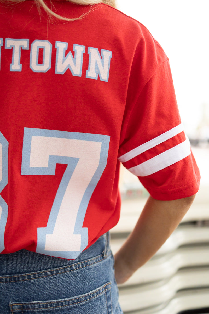 Girl wearing red vintage inspired baseball tee with white stripes on the short sleeves, vintage font spelling "Maverick" on the front and "Cowtown '87" on the back