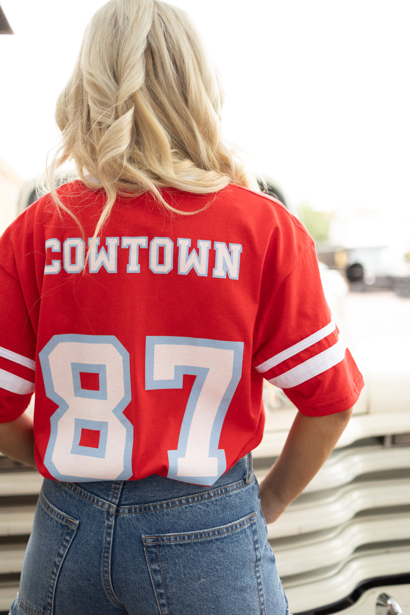 Girl wearing red vintage inspired baseball tee with white stripes on the short sleeves, vintage font spelling "Maverick" on the front and "Cowtown '87" on the back