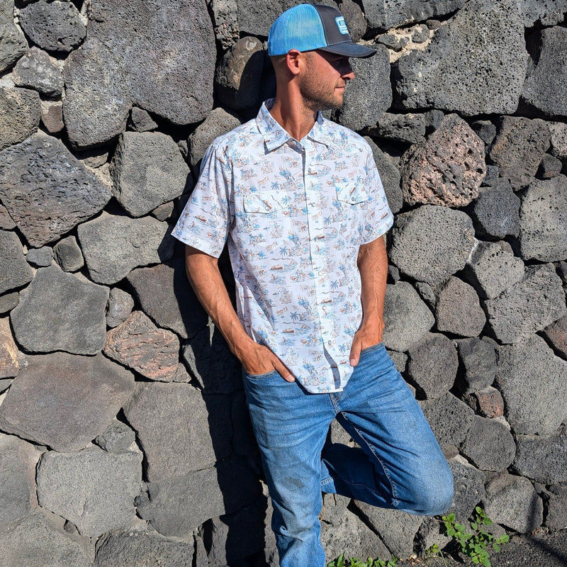 Man wearing a patterned shirt and blue jeans standing against a stone wall.