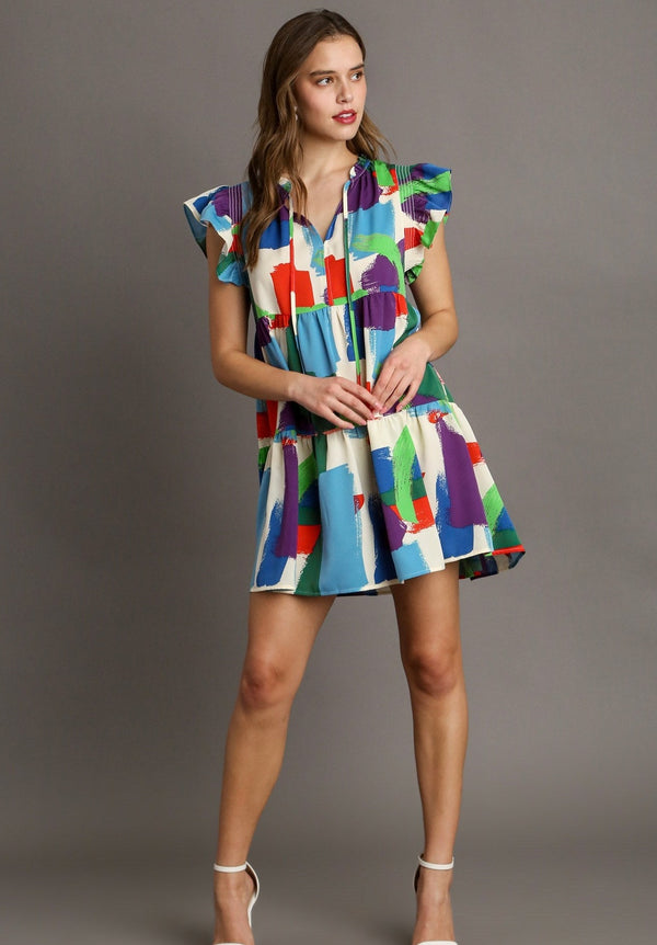 Woman wearing tiered mini dress with ruffle sleeve, brush stroke pattern and decorative strings by the neckline