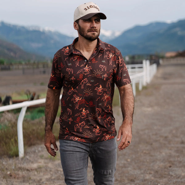 Man wearing a patterned shirt and cap standing in an outdoor setting with mountains in the background