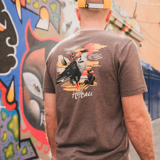 Heather brown t-shirt with graphic design on the back. Graphic design containg the image of a bird looking to its left with a snake in its mouth with the scenery of a desert in a orange, yellow, white and black color way. The bottom of the shirt contains the words "Heed the Call".