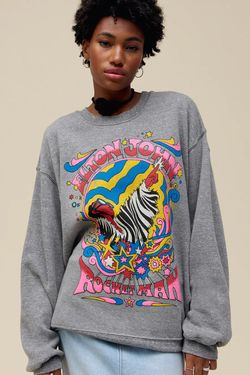 Woman wearing grey long sleeve top with vibrant colors printing "Elton John" And Graphic of person pointing upward