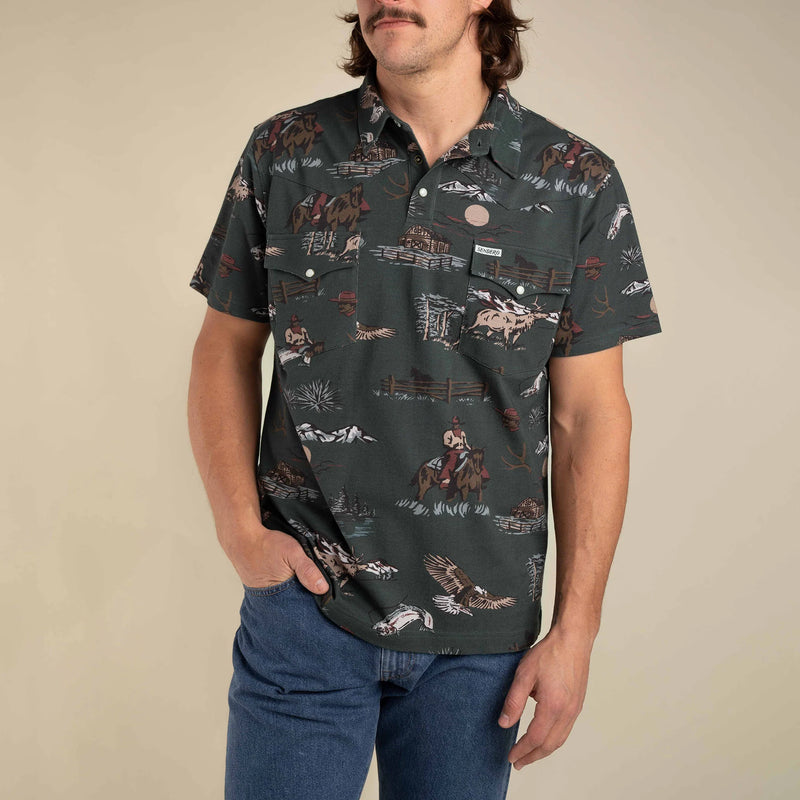 Dark green short sleeve polo shirt with double breast pockets with cartoon images of cowboy on horse, fence, fish, mountains, elk, eagle, and sun all repeated throughout the shirt