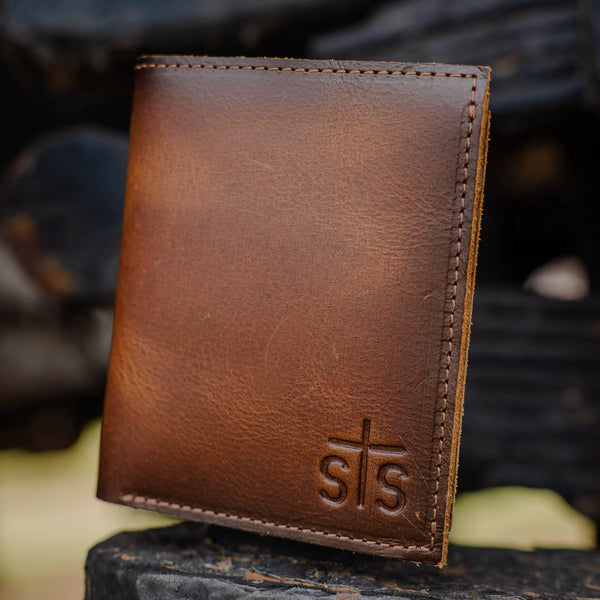 Brown leather wallet with 'sts' logo on a blurred natural background