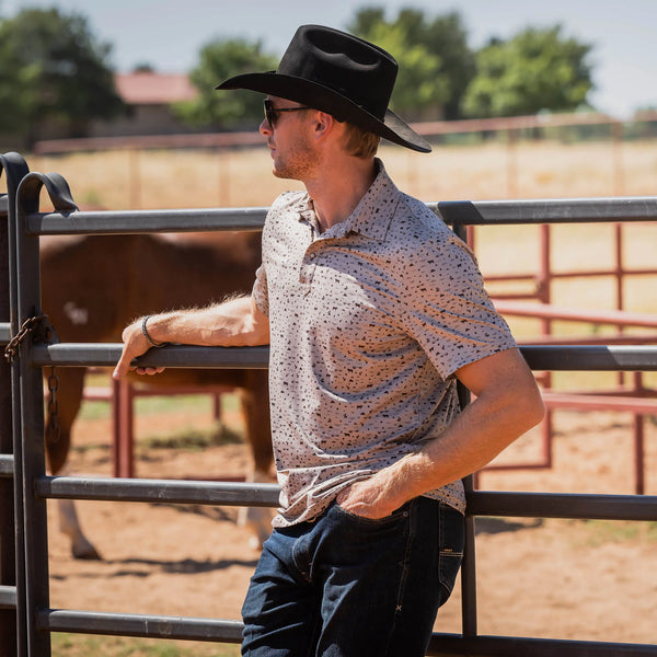 Man wearing a cowboy hat and patterned shirt standing next to a metal fence with a horse in the background.