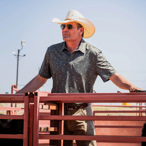 Man wearing a cowboy hat and sunglasses standing behind a red metal fence.