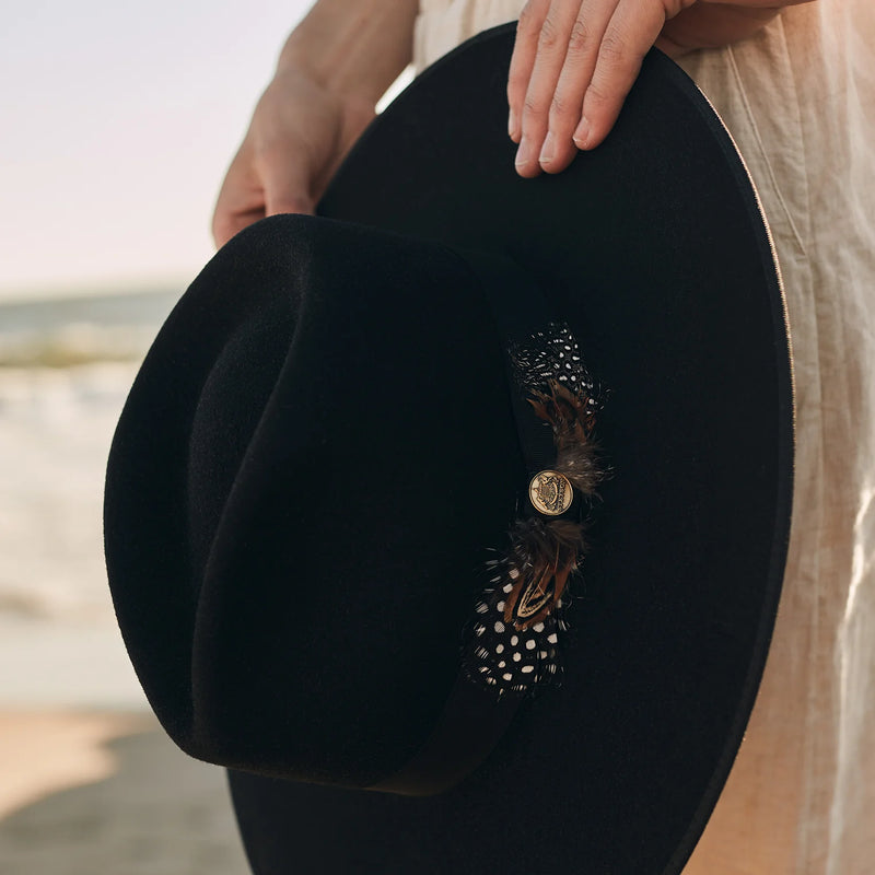 Black hat with decorative feather and button held by a person against a blurred background