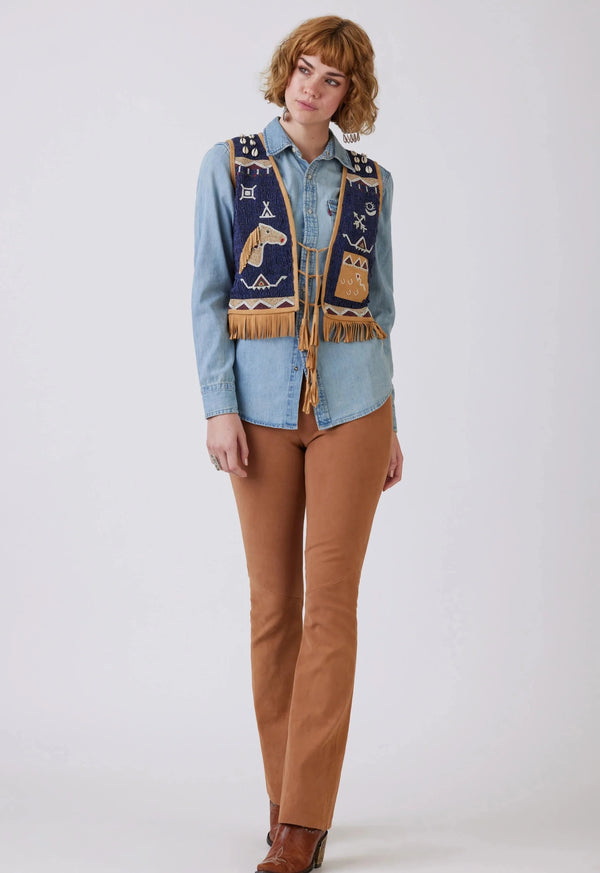 Woman wearing goat suede vest, completely covered in intricate hand-beading depicting classic pictorials of the Plains and Southwestern shapes and symbols, complemented by cowrie shells and finished with fringe and a 3-tie tassel closure.