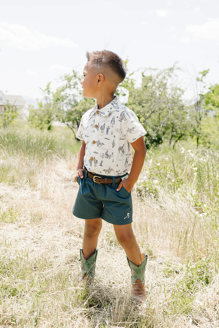 Child wearing a patterned shirt, shorts, and boots standing in a grassy field.