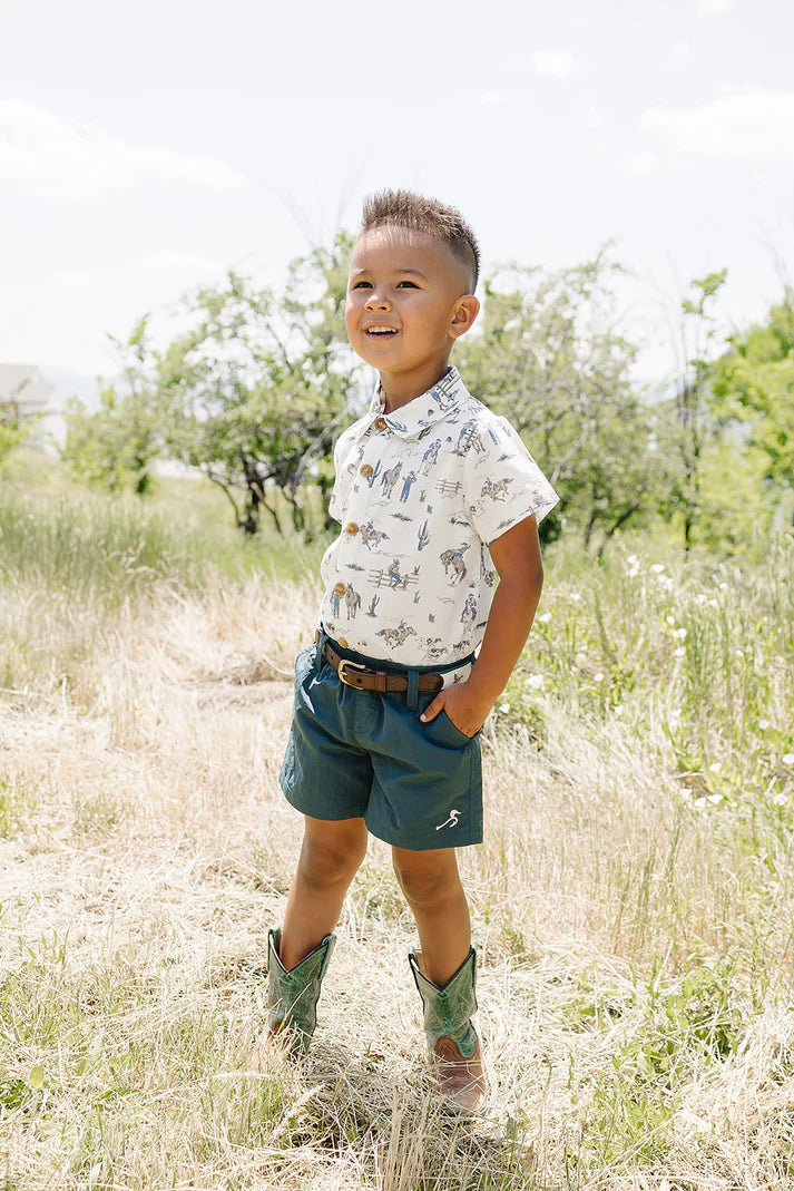 Child in a field wearing a patterned shirt and shorts with cowboy boots.