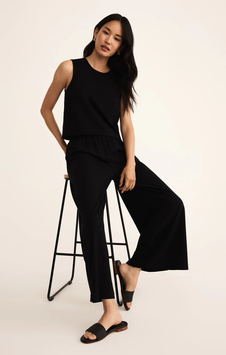 Woman wearing a black sleeveless top and wide-leg pants sitting on a stool against a white background
