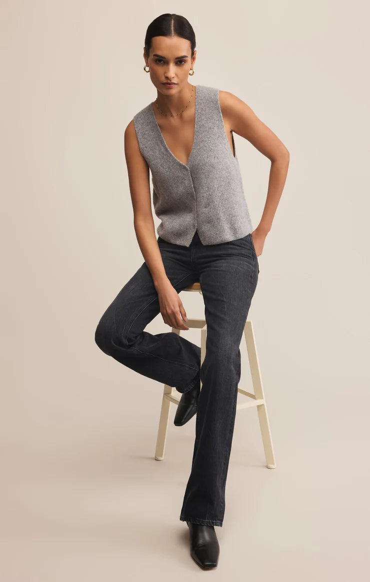 Woman wearing a gray sleeveless sweater vest and dark jeans sitting on a stool against a beige background