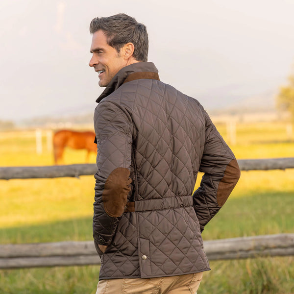 Man wearing a quilted jacket with brown elbow patches in a field with a horse.