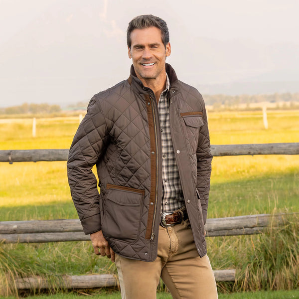 Man wearing a quilted jacket standing in a field with a wooden fence.