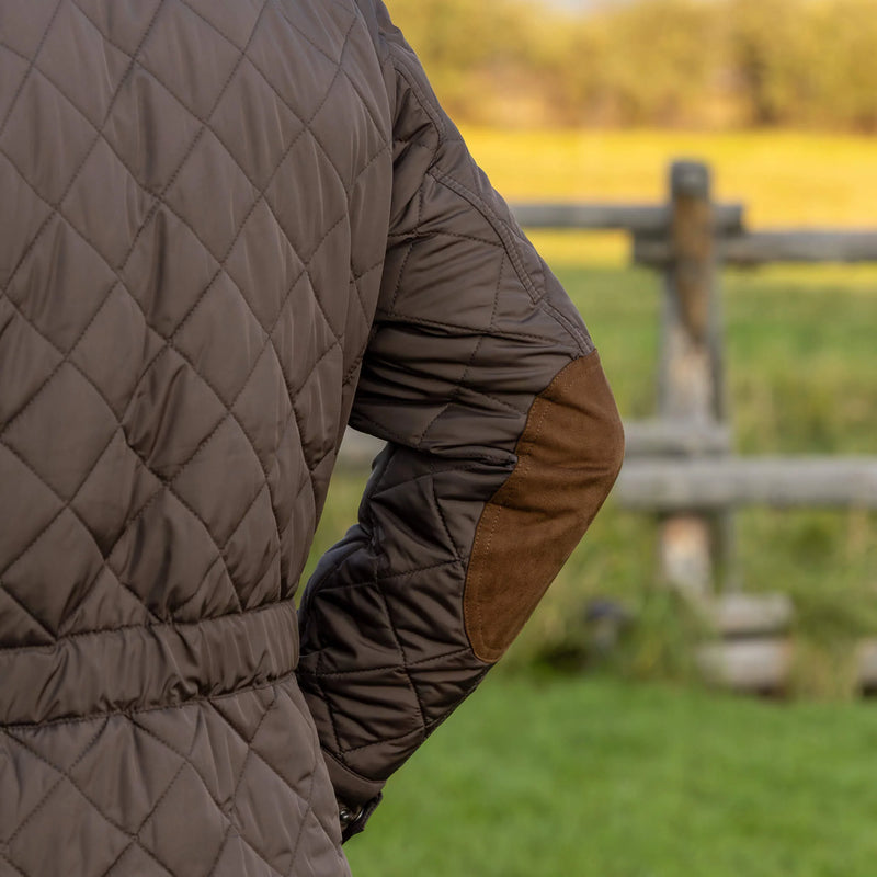 Person wearing a quilted brown jacket in a field with a wooden fence in the background