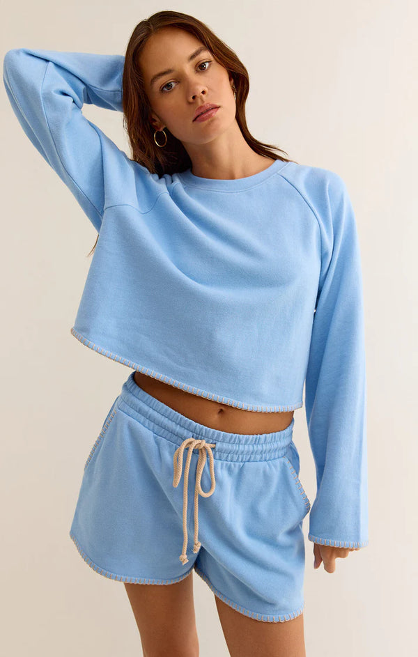 Woman wearing baby blue long sleeve pullover with cream embroidery detail on the hem and the cuffs of the sleeves 