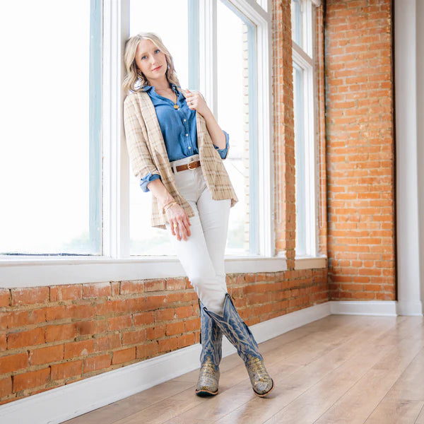 Woman standing in a room with brick walls and large windows, wearing a blue shirt, beige cardigan, white pants, and blue boots.
