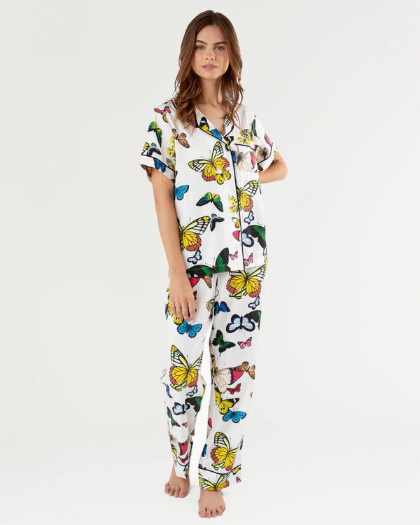 Woman wearing white background pajamas with vibrant, multicolor butterflies all over. The top is a short sleeve and the bottoms are long pants